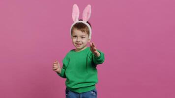 linda pequeño chico vistiendo mullido conejito orejas en estudio y ondulación, siendo adorable en contra rosado antecedentes. sonriente joven niño siendo emocionado acerca de Pascua de Resurrección fiesta festividad, infancia inocencia. cámara una. foto