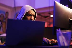 Focused african american scammer cracking password to gain access to system. Concentrated hacker in hood hacking database and breaching data in dark hideout place with neon light photo