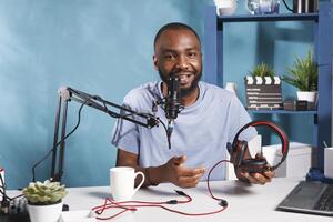 sonriente Internet hombre de influencia promoviendo auriculares mientras grabación y mirando a cámara. alegre africano americano vlogger revisando dispositivo, Hablando en micrófono y radiodifusión En Vivo foto