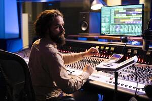 Music technician mixing and mastering songs with motorized faders and knobs, using control desk dashboard and computer. Young sound engineer creating tracks for an album in studio. photo