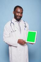 Portrait of black man wearing a stethoscope and lab coat is holding a tablet with green screen. African american doctor grasping device displaying blank chromakey mockup template. photo