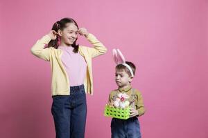 Little kids posing with festive handmade decorations for easter celebration, feeling excited about spring holiday tradition. Sweet brother and sister showing painted eggs in a basket. photo