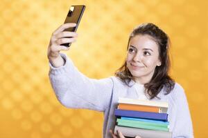 Smiling woman doing selfie with cellphone holding stack of books, isolated over studio background. Happy student with pile of textbooks in arms taking picture with mobile phone photo