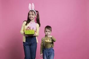 Young kids holding handmade decorations and ornaments on camera, posing for an easter celebration photoshoot. Adorable boy and girl feeling joyful about spring celebration event in studio. photo