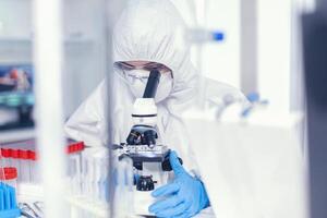 Scientist in sterile protection suit analysing coronavirus looking through microscope. Chemist in coverall examining vaccine evolution using high tech researching diagnosis against covid19 virus photo