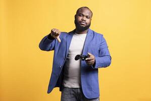 Upset african american man showing thumbs down sign gesturing holding controller, disapproving of games. Disappointed BIPOC person against gaming, doing rejection hand gesture, studio background photo