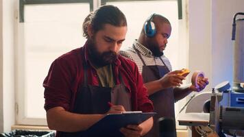 artesano dibujo planos en bloc en estudio con bipoc compañero de trabajo en antecedentes resolviendo tareas. fabricante mirando a técnico esquemas a ejecutar carpintería proyectos, cámara un foto