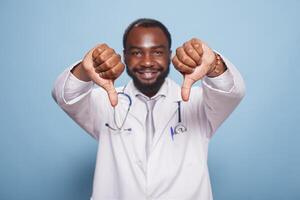 Black man wearing a hospital uniform and stethoscope standing and doing thumbs down to the camera. Portrait of african american medical specialist making disapproval hand gestures. photo