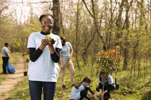 Young teenager activist planting small green seedlings in the woods, restoring natural habitat and protecting the environment. Proud african american girl doing voluntary work to save the planet. photo