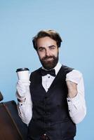 Confident doorman feeling happy in studio, holding cup of coffee against blue background. Smiling bellhop employee wearing classy attire and drinking cold brew of tea refreshment. photo