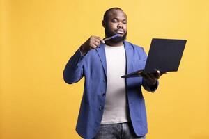 African american man composing email, typing on laptop keyboard, struggling to think of what to write. BIPOC person sending mail online, brainstorming message idea, studio background photo