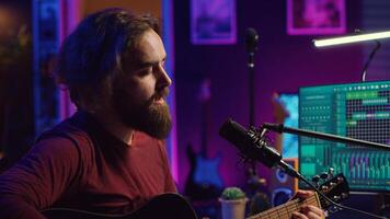 Songwriter producing tunes on acoustic guitar in his personal studio, using a microphone to sing the chords. Audio engineer works with soundboard and equalizer to compose new music. Camera A. photo