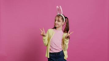 Young cheerful kid with pigtails showing peace sign in studio, feeling excited and confident about easter sunday celebration. Smiling youngster does signs against background. Camera B. photo