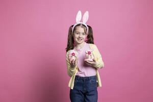 joven linda niño con conejito orejas demostración rosado Pascua de Resurrección adornos en frente de cámara, participación su hecho a mano huevo y Conejo juguete. pequeño niño sonriente en estudio y celebrando primavera día festivo. foto