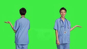 Medical assistant pointing at something aside and doing web commercial, creating advertisement against greenscreen backdrop. Young nurse with scrubs showing a thing to left or right sides. Camera B. photo