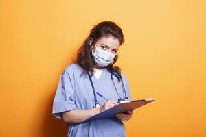 mujer médico en azul matorrales tomando notas en portapapeles archivos, en pie en contra aislado antecedentes. cuidado de la salud especialista con cara máscara escritura en médico documentos, mirando a cámara. foto