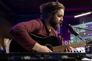 Songwriter composing a new song on acoustic guitar in home studio, using a microphone to sing the chords. Artist works with mixing console and daw software, guitarist producing new music. photo