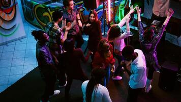 Multiethnic group of friends partying, showing funky dance moves at nightclub. Men and women dancing on electronic beats having fun on night out, enjoying colorful spotlights. Handheld shot. photo