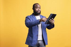 Bored person mindlessly scrolling on tablet screen, isolated over studio background. African american man passing time on social media on portable device, enjoying leisure time photo