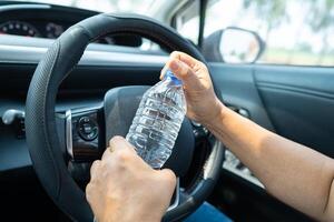 conductora asiática sosteniendo una botella para beber agua mientras conduce un automóvil. una botella de agua caliente de plástico provoca un incendio. foto