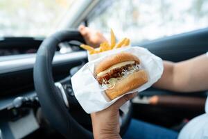 asiático dama participación hamburguesa y francés papas fritas a comer en auto, peligroso y riesgo un accidente. foto