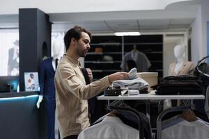 Young man shopping for trendy menswear, browsing through shelf in clothing store. Fashion boutique customer choosing apparel and accessory, searching for size and examining style photo