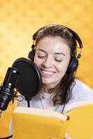 Happy narrator wearing headphones reading aloud from book into mic against yellow background. Joyous lady recording audiobook, creating engaging media content for listeners photo