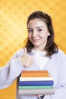 retrato de alegre mujer que lleva apilar de libros, reunión información para colegio examen, demostración pulgares arriba, estudio antecedentes. contento estudiante participación libros de texto, haciendo positivo mano gesto foto