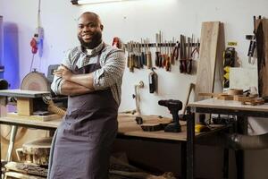 retrato de sonriente africano americano carpintero preparando a comienzo producción en sucio mueble montaje tienda. contento bipoc fabricante a banco de trabajo Listo a cortar madera piezas foto