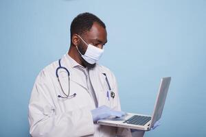 Medical physician with latex gloves and face mask holding a laptop to review findings of a clinical diagnosis. Portable computer is being held by medic wearing lab coat and stethoscope. photo