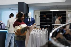 sonriente africano americano mujer participación chaqueta en percha mientras compras para atuendo en ropa almacenar. Moda boutique cliente explorador vestir estante y seleccionando formal vestir foto