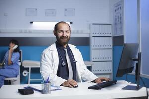 Middle aged doctor smiling at his desk in a medical office. Caucasian male physician appears to be wearing a white lab coat, female nurse is in the background on a call. Portrait shot. photo