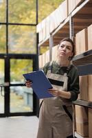 Stockroom employee supervising cardboard packages in mail sorting center and writing inventory clipboard. Post office warehouse manager searching customer parcel, checking postal form. photo