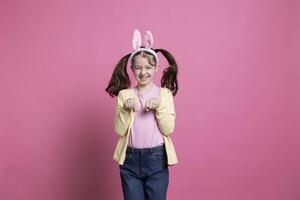adorable pequeño niña gasto Pascua de Resurrección fiesta por saltando alrededor me gusta un Conejo para el cámara mientras vistiendo conejito orejas. Encantado contento joven jugando y riéndose en rosado antecedentes. foto