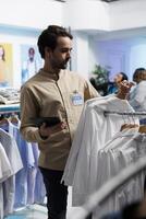 ropa Tienda asistente comprobación inventario con digital tableta y mostrando vestir en estante. Moda boutique gerente colgando prenda a atraer clientes en compras centrar foto