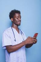 africano americano médico en matorrales usa estetoscopio y sostiene un teléfono inteligente, sonriente con confianza en un estudio. negro moderno cuidado de la salud profesional utilizando móvil dispositivo durante codicioso 19 pandemia. foto