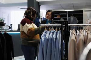 Clothing store woman asian assistant talking and consulting customer. Customer exploring rack with apparel, choosing formal outfit and getting help from boutique employee photo