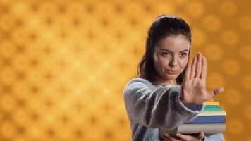 Portrait of stern woman holding stack of books doing stop sign gesturing, studio background. Student with pile of textbooks in arms used for academic learning doing halt hand gesture, camera A photo