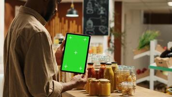 African american client holding tablet with greenscreen layout in local organic shop, looking at chromakey next to additives free goods. Regular customer uses device with isolated copyspace. Camera 1. photo