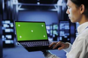 IT consultant with green screen laptop between server hub clusters providing processing resources for businesses worldwide. Worker uses mockup device to fix data center mainframes photo