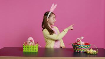 Adorable little kid indicating all directions in front of camera, pointing up, down, left and right while sitting at the table. Smiling lovely girl with bunny ears looking around. Camera B. photo