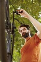 Detailed outside image shows youthful healthy caucasian man inspecting and maintaining bicycle components. Dedicated male cyclist outdoors repairing bike crank arm and pedal for summer cycling. photo