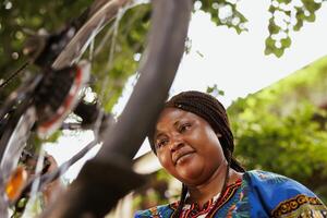 Young sports-loving black woman examining bicycle tire in home yard for damage to repair with tools. Energetic african american female fixing broken bike chain and wheel outside. photo