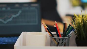 Focus on desk belongings in modern office with notebook in blurry background showing business diagram and figures, close up. Pencil holder, mini house plant, empty coffee mug and graphs on laptop photo