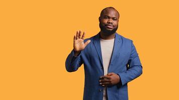 African american man reluctantly doing salutation hand gesture, saying goodbye. Portrait of sad BIPOC person raising arm to greet someone after leaving, isolated over studio background, camera B photo