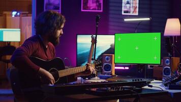 Young artist playing his guitar and practicing new song with online tutorial, looks at computer with greenscreen display. Musician learning new acoustic instrument accords, home studio. Camera A. photo