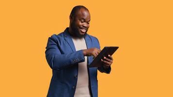 Smiling man scrolling on tablet touchscreen, using internet and credit card to pay for products. African american person happily browsing online shopping websites, studio background, camera B photo