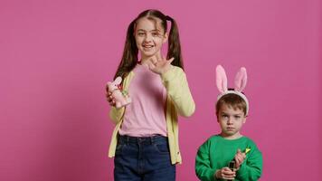 Cute brother and sister posing against pink background in studio, wearing bunny ears and playing with toys. Cheerful siblings feeling excited about easter, traditional spring holiday. Camera B. photo