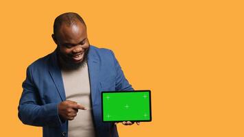 Upbeat african american man holding green screen tablet, doing recommendation. Cheerful BIPOC person pointing towards mockup device, giving positive feedback, studio background, camera A photo