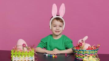 Young small kid placing bunny ears on his head in studio, preparing for easter sunday holiday celebration. Cheerful little boy sitting at a table to paint eggs and craft festive ornaments. Camera A. photo